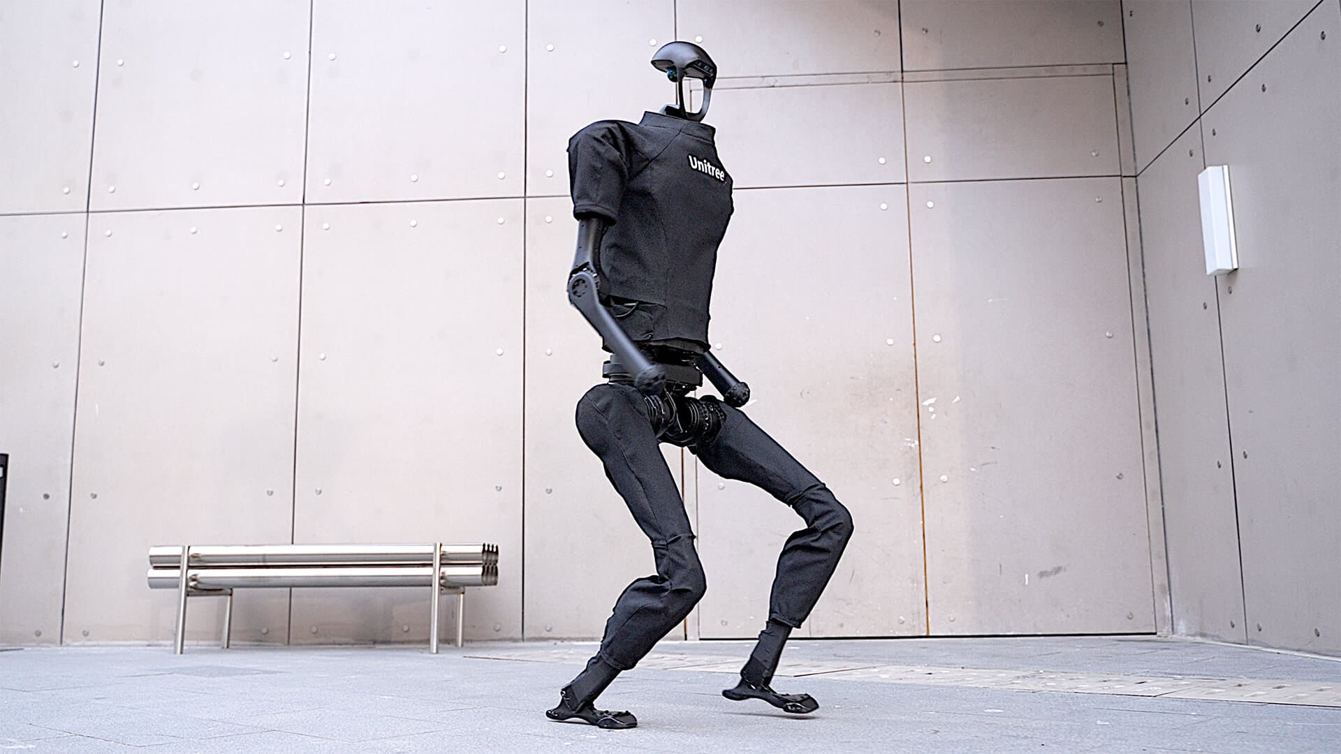 Unitree H1 humanoid robot mid-stride in front of a concrete wall
