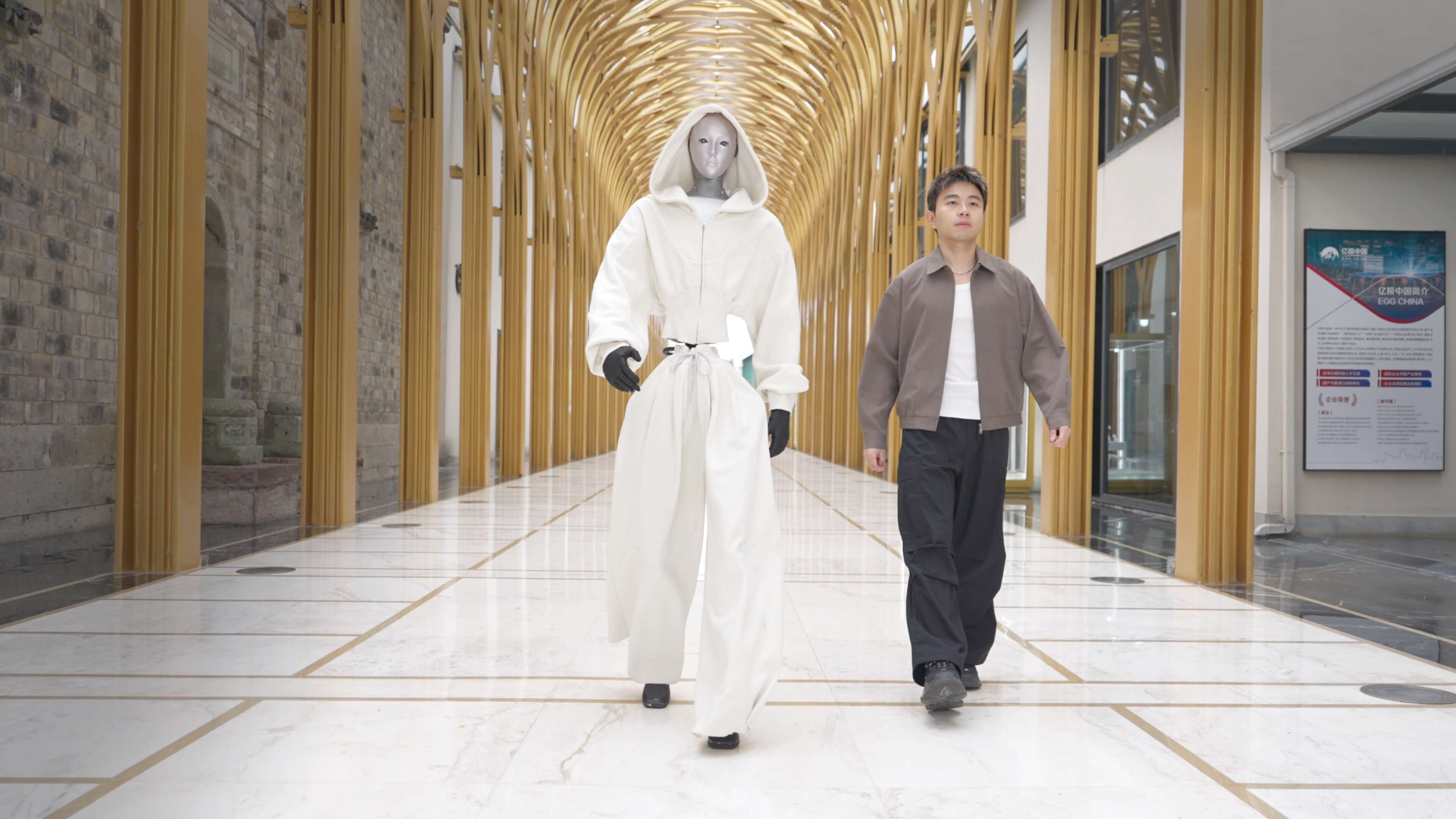 Unitree H2 walking beside a man in a corridor, both roughly the same height